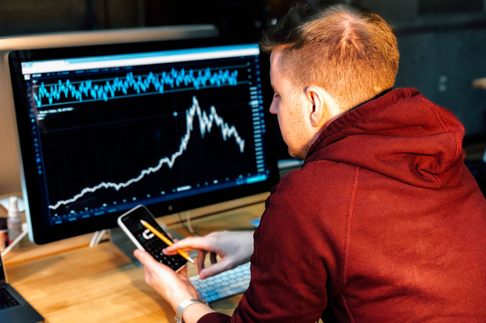 Mann mit Taschenrechner vor Monitor mit Börsenchart auf dem Bildschirm